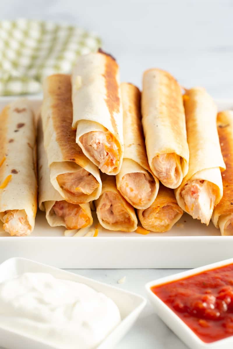 Taquitos stacked on serving trap with sour cream and salsa in dipping dishes
