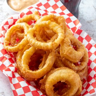 Finished onion rings in serving basket with pop and sauce in background