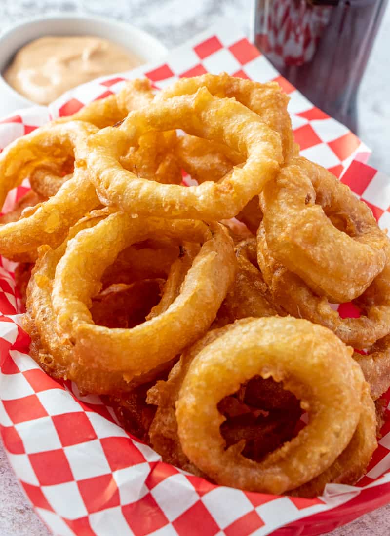 Onion rings in bar food basket stacked on top of one another with dipping sauce in background