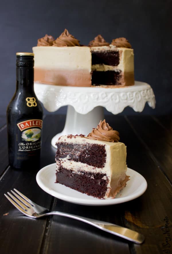 Slice of cake on plate with whole cake on cake stand next to bottle of Baileys