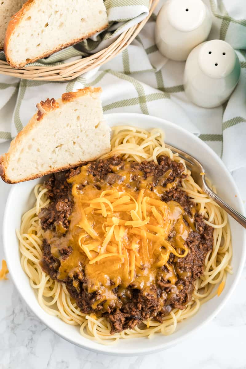 Chili plated in bowl with melted cheese and slice of bread with bread basket on side