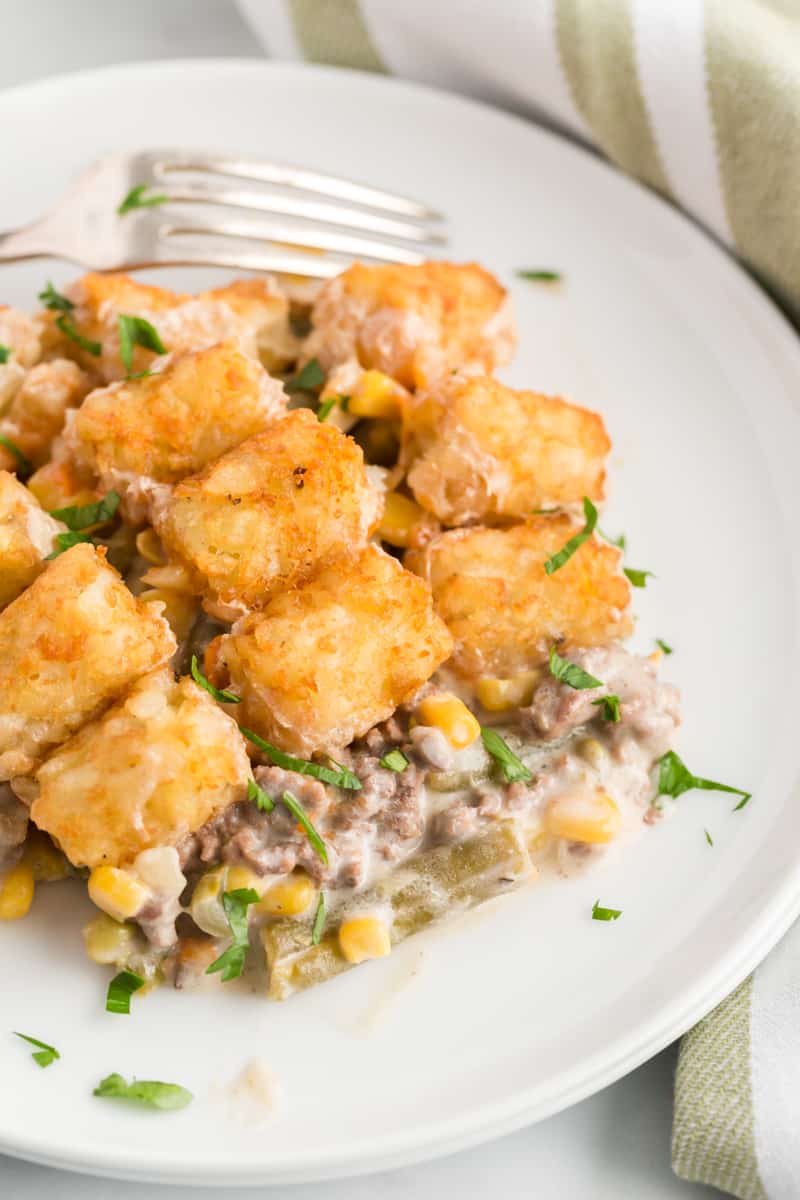 Tater tot hotdish on plate with fork with browned tater tots on top of filling sprinkled with a little parsley