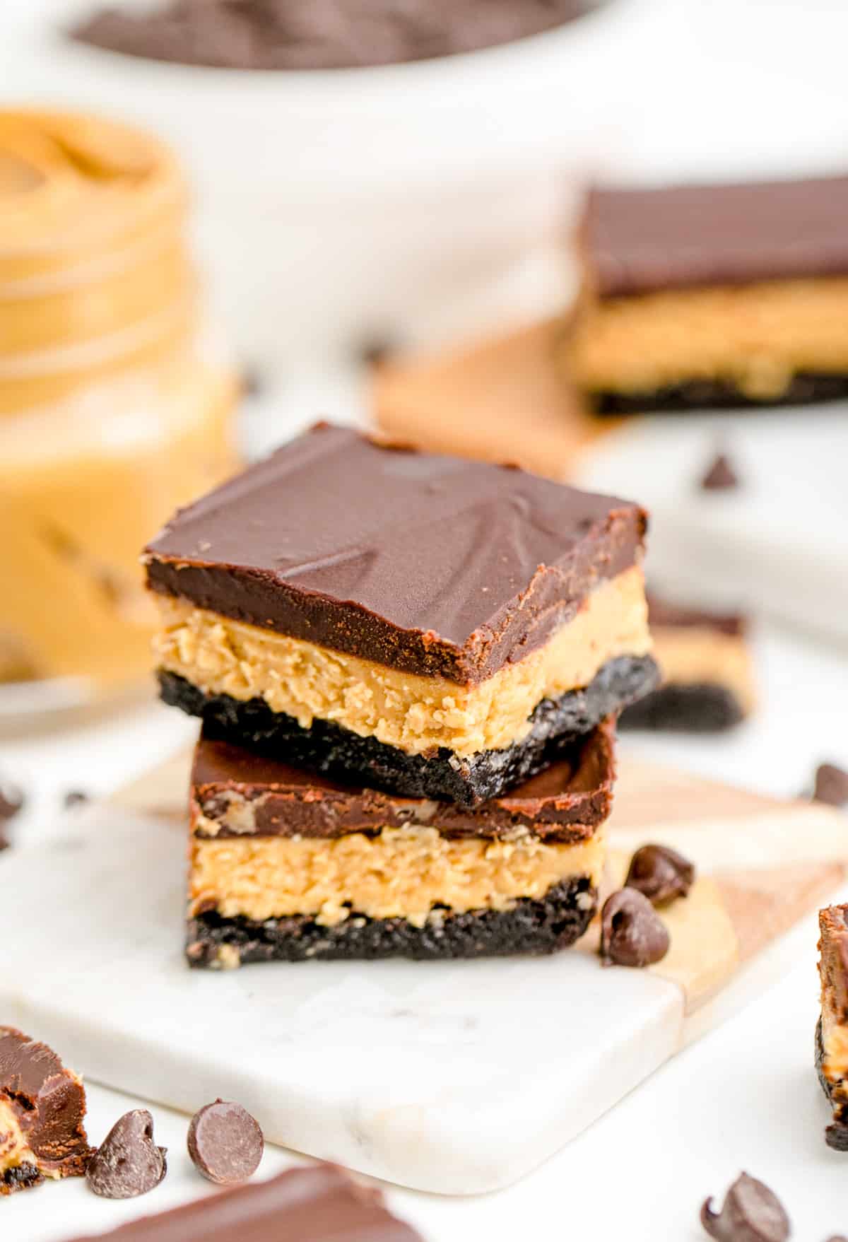 Two stacked Ultimate Peanut Butter Brownies on marble square with chocolate chips next to them and peanut butter in background.