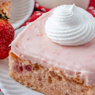 Slice of strawberry cake on white plate topped with a swirl of whipped cream