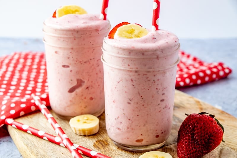 Two Strawberry Banana Smoothies with red and white straws on brown board