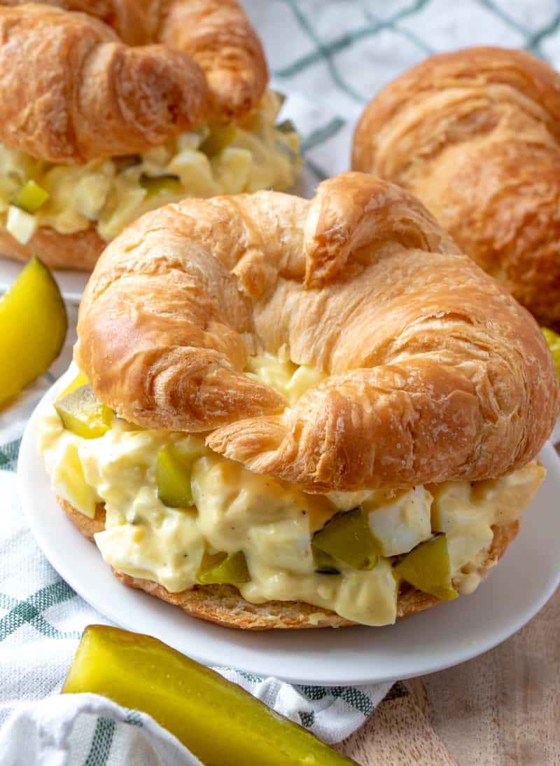 Egg Salad Recipe on croissant on plate with pickles around it