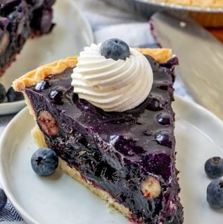 Slice of blueberry pie on white plate topped with whipped cream and a blueberry