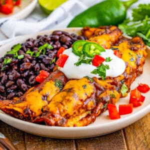 Square image of two Enchiladas on white plate with garnishes and served with black beans.