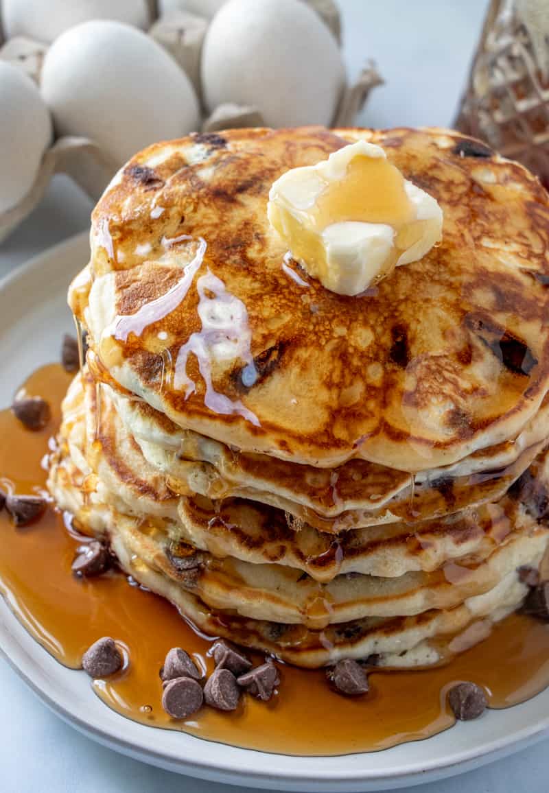 stacked chocolate chip pancakes with pat of butter, syrup over top running down sides and chocolate chips on plate