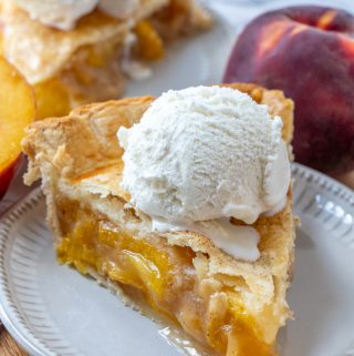 Slice of finished peach pie on white plate topped with vanilla ice cream