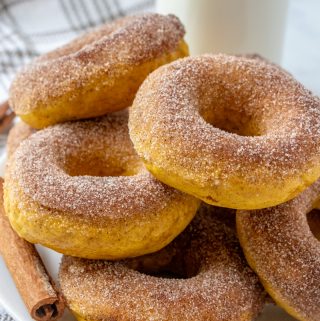Donuts stacked on white round plate with cinnamon stick on left side and milk in background
