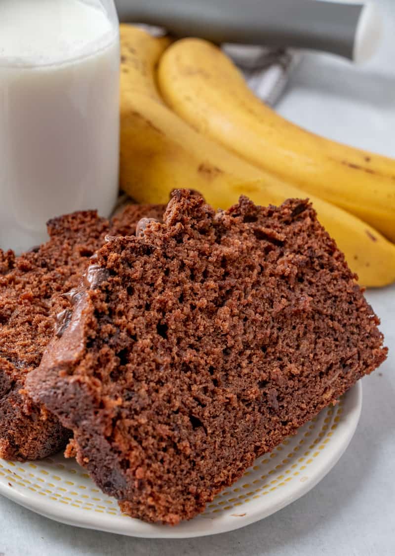 Two slices of chocolate banana bread on plate with bananas in background