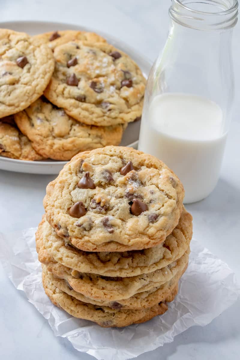 Cookies stacked on top of one another with a plate of cookies in background with milk