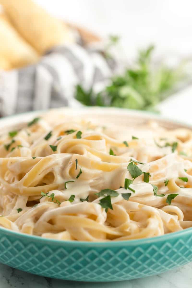 Close up photo of Alfredo sauce served over pasta I bowl with parsley and breadsticks in background