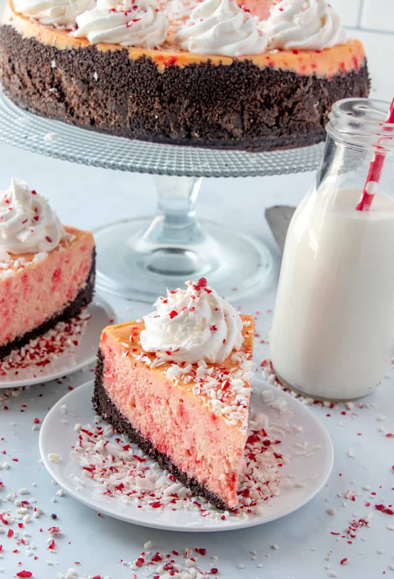 Slice of cheesecake on plate with whole cheesecake on cake stand in background and glass of milk