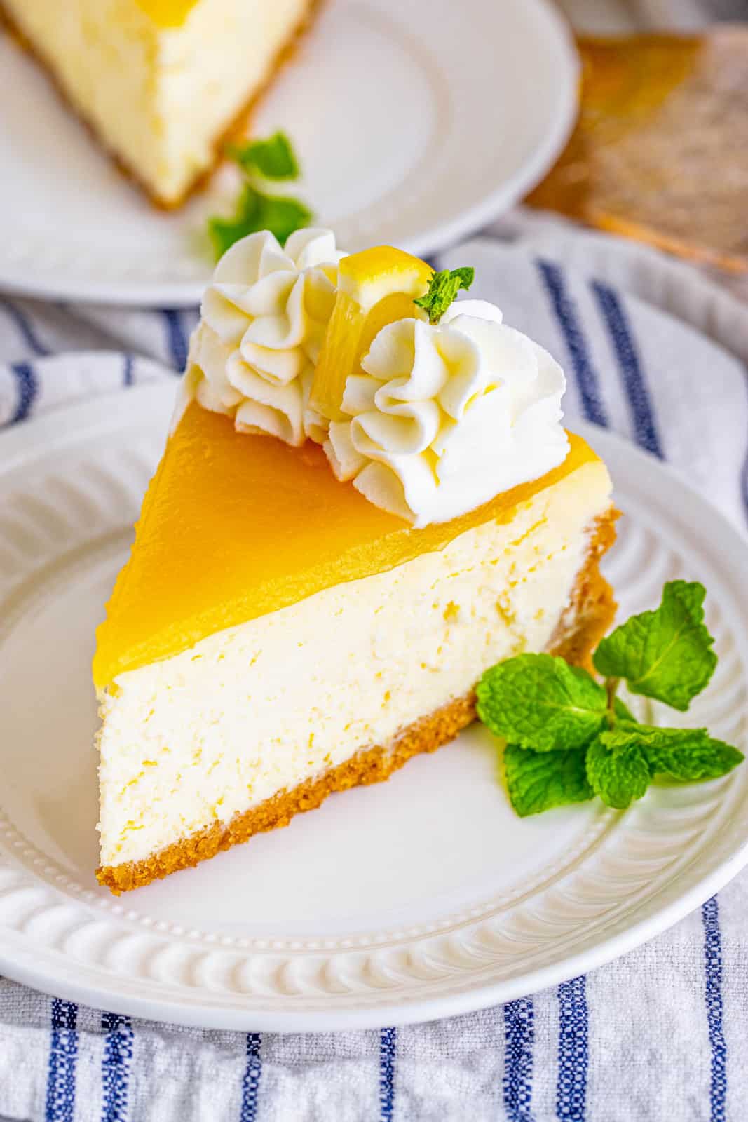 Slice of Lemon Cheesecake on white plate with whipped cream, lemon and mint.