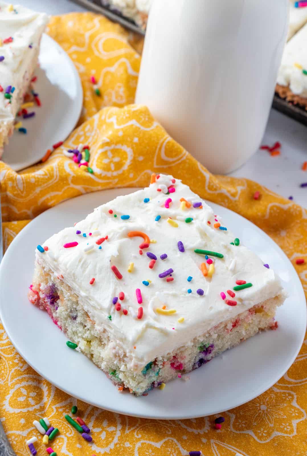 Slice of Funfetti Cake on white plate topped with sprinkles and milk in background