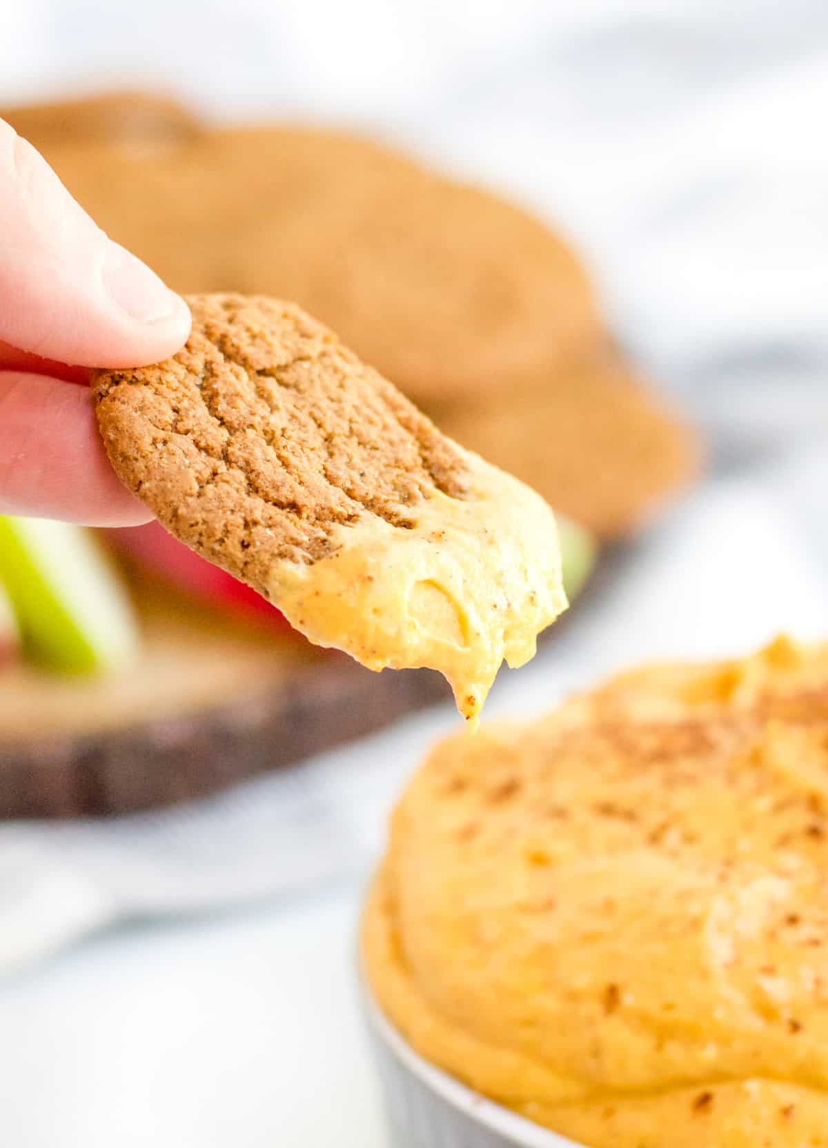 Hand holding a cookie with dip on it with dip in bowl and background along with other dippers.