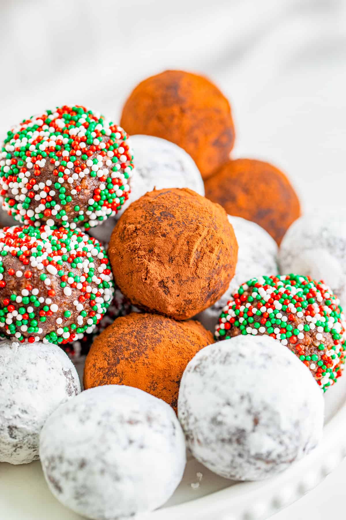 Finished Rum Balls rolled in powdered sugar, sprinkles and cocoa powder on white cake stand.