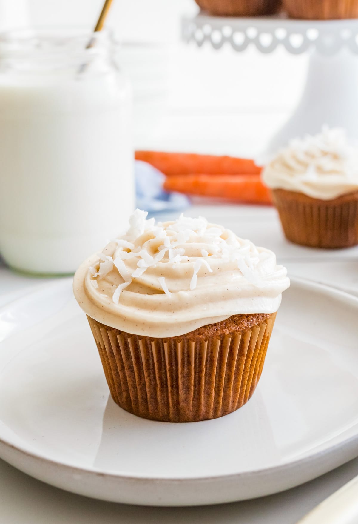 One cupcake on white plate with milk and carrots in the background.