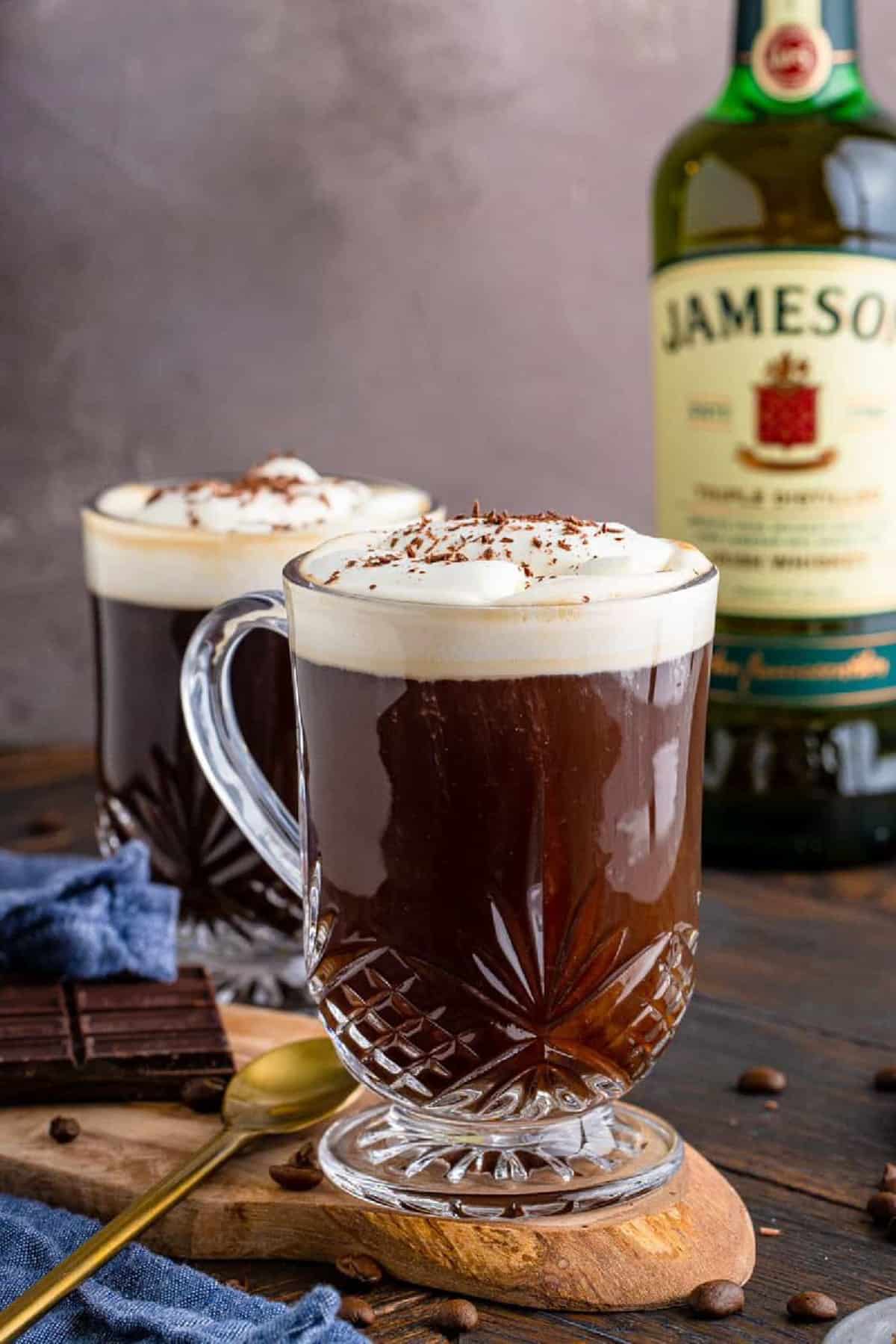 Irish Coffee Recipe on wooden board with whiskey in background and coffee topped with whipped cream and chocolate shavings.