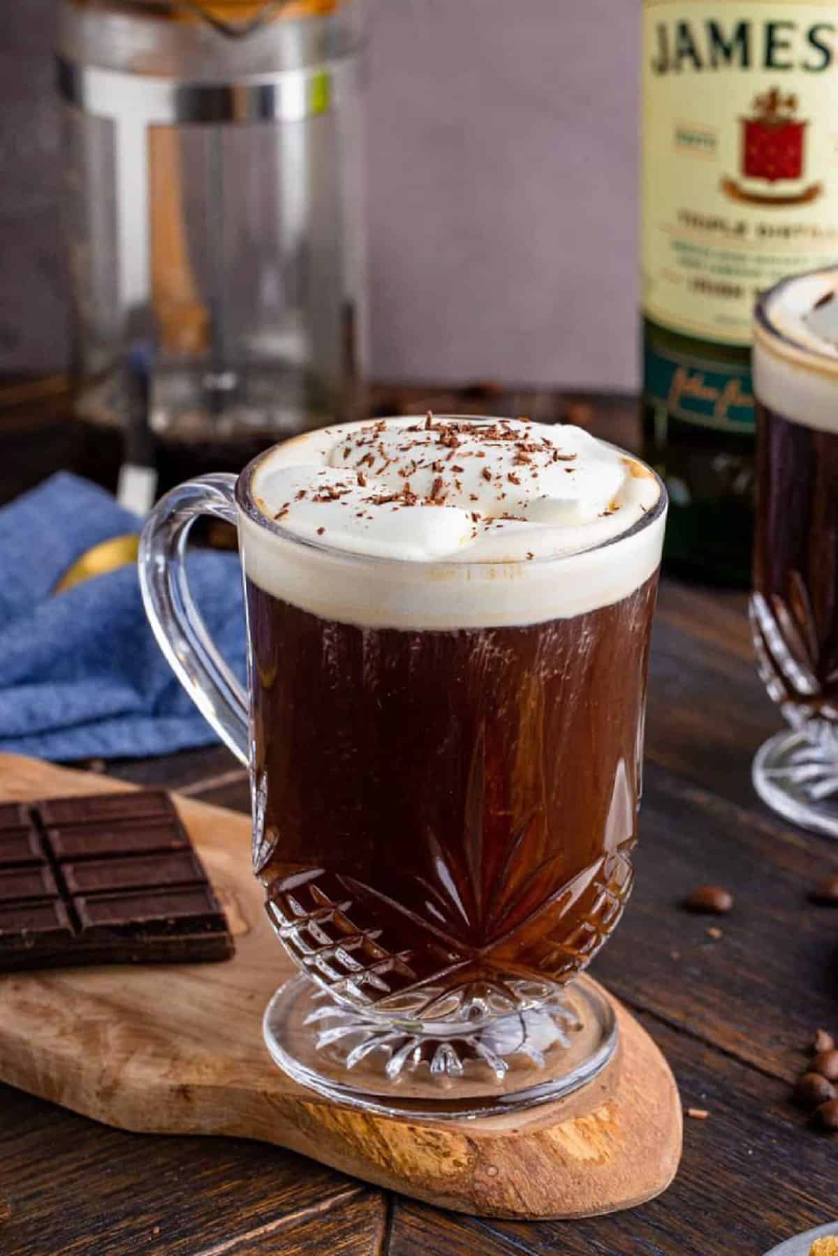 One coffee on wooden board with french press and whiskey in background topped with whipped cream and chocolate shavings.