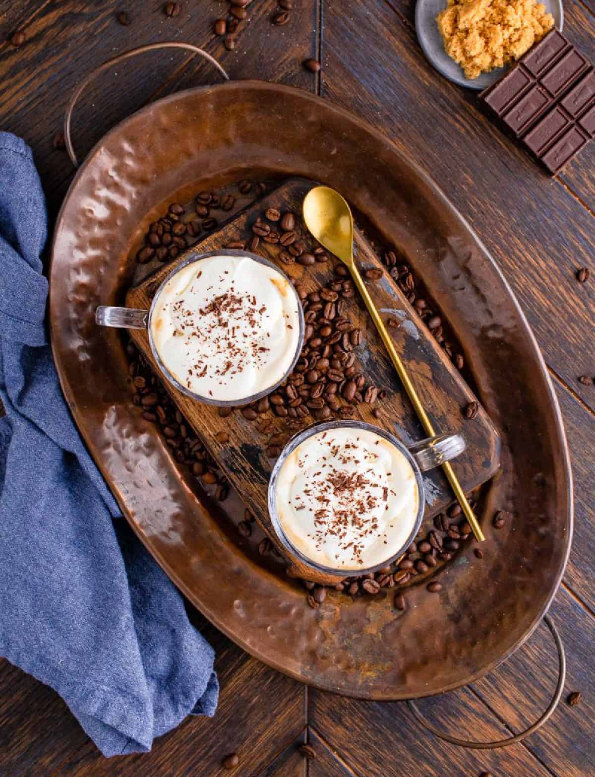 Overhead of two coffees on bronze metal serving tray with long golden spoons topped with whipped cream and chocolate shavings.