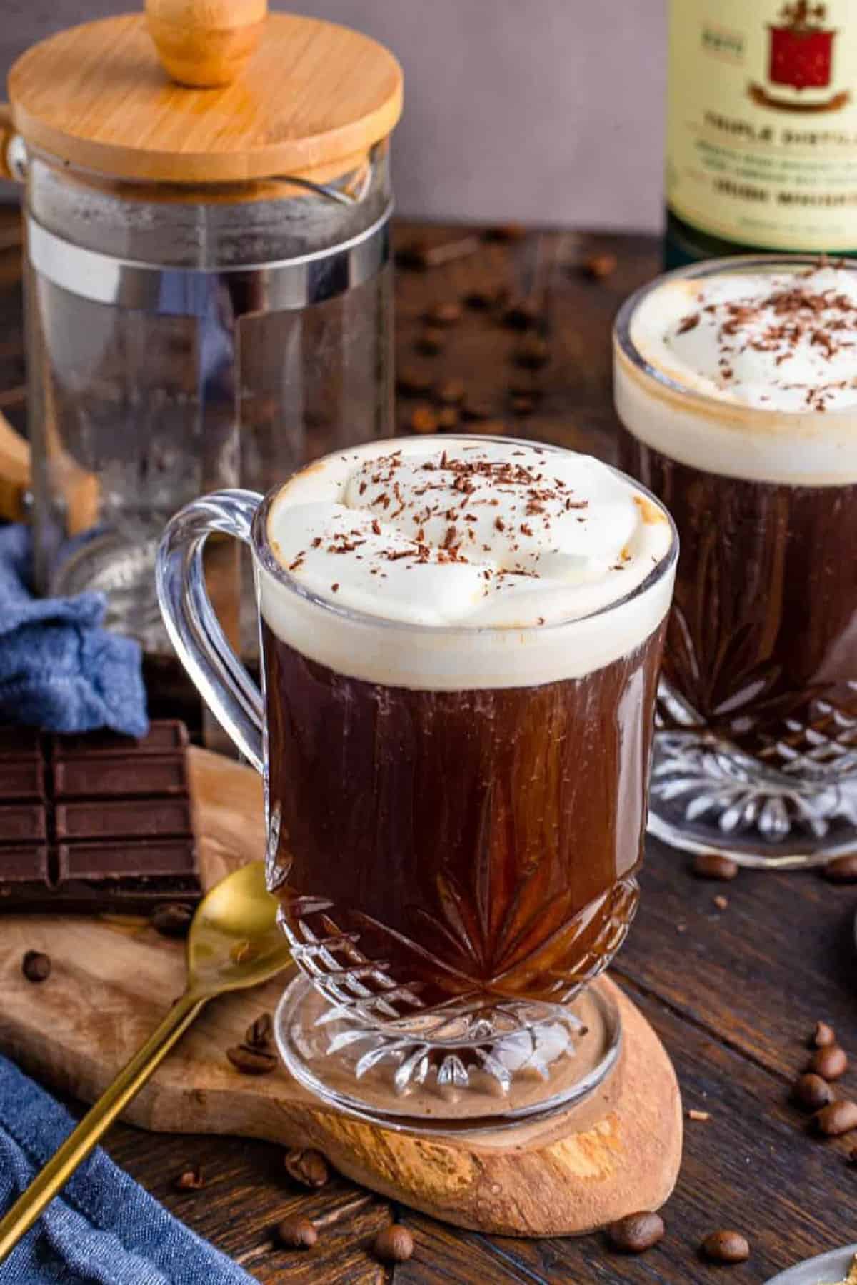 Irish Coffee Recipe on wooden board with long golden spoon with french press in background and topped with whipped cream and chocolate shavings.