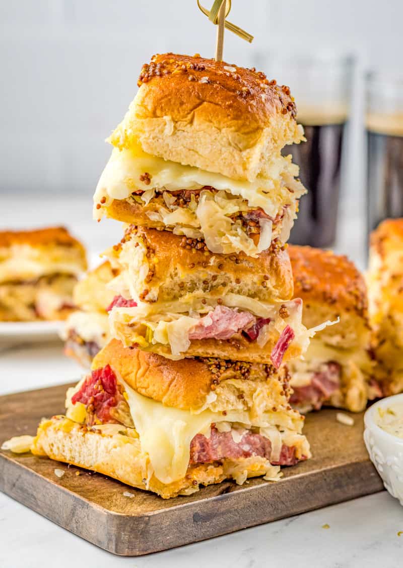 Three stacked Reuben Sliders on wooden board showing melted cheese and other layered ingredients.