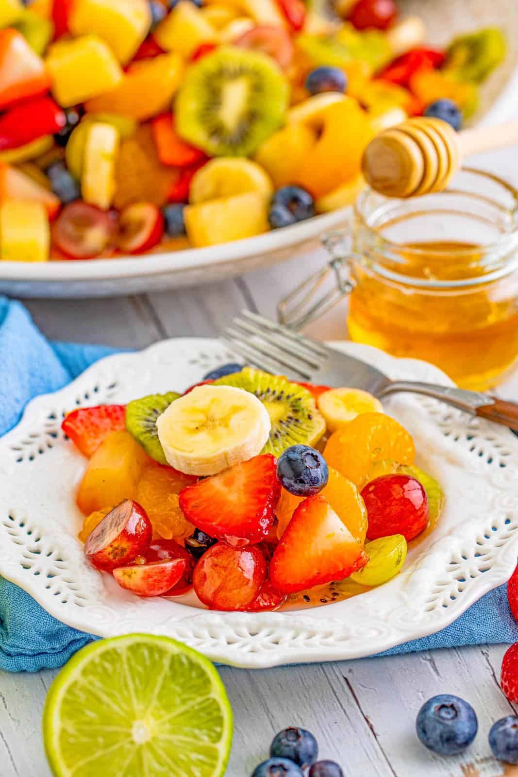Plate of Fruit Salad with honey and large bowl of salad in background.