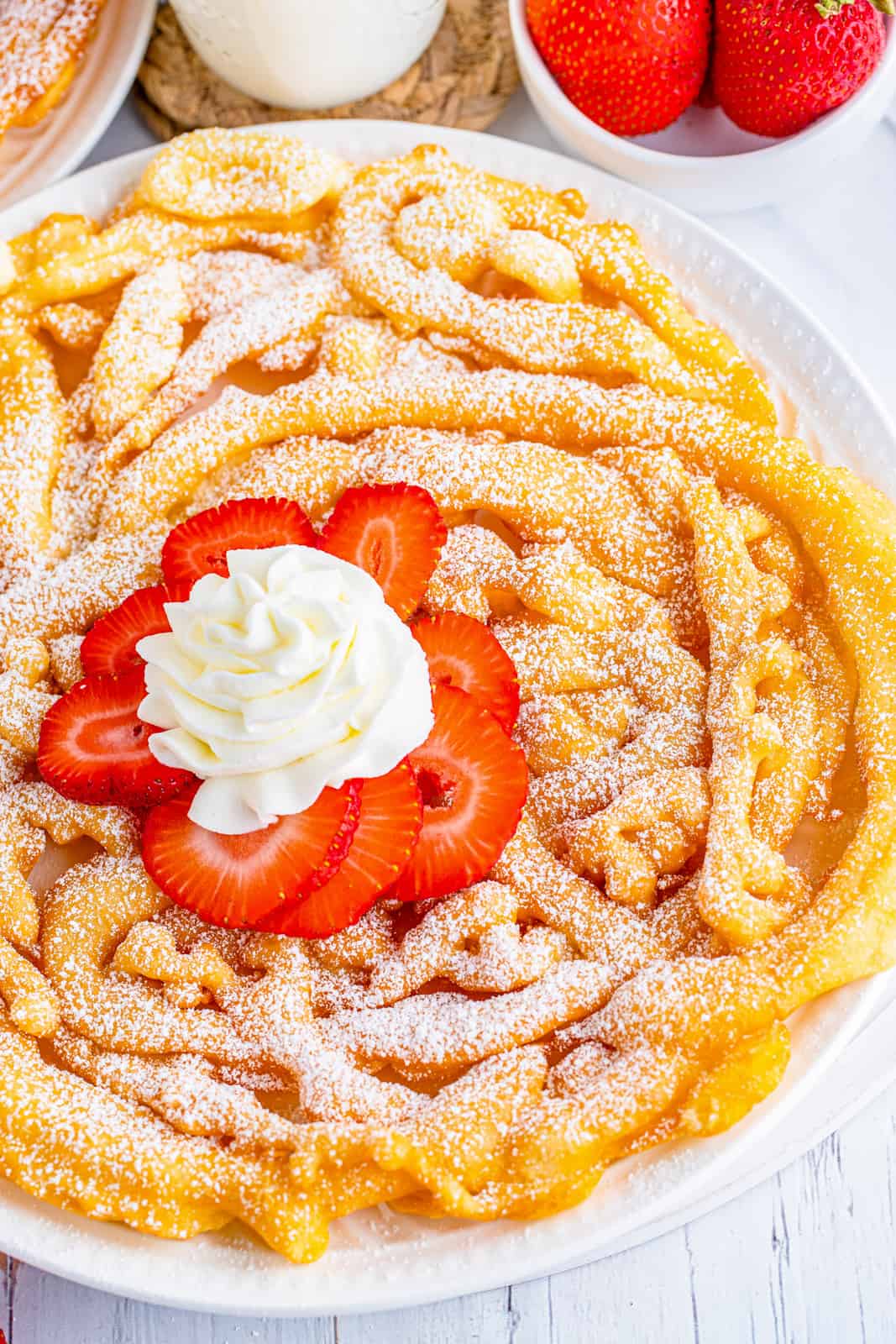 Overhead photo of finished Funnel Cake on white plate topped with powdered sugar, strawberries and whipped cream