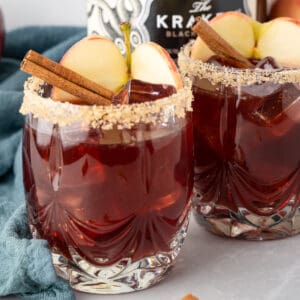 Close up square image of two glasses of sangria garnished with brown sugar rim, apple and cinnamon stick.
