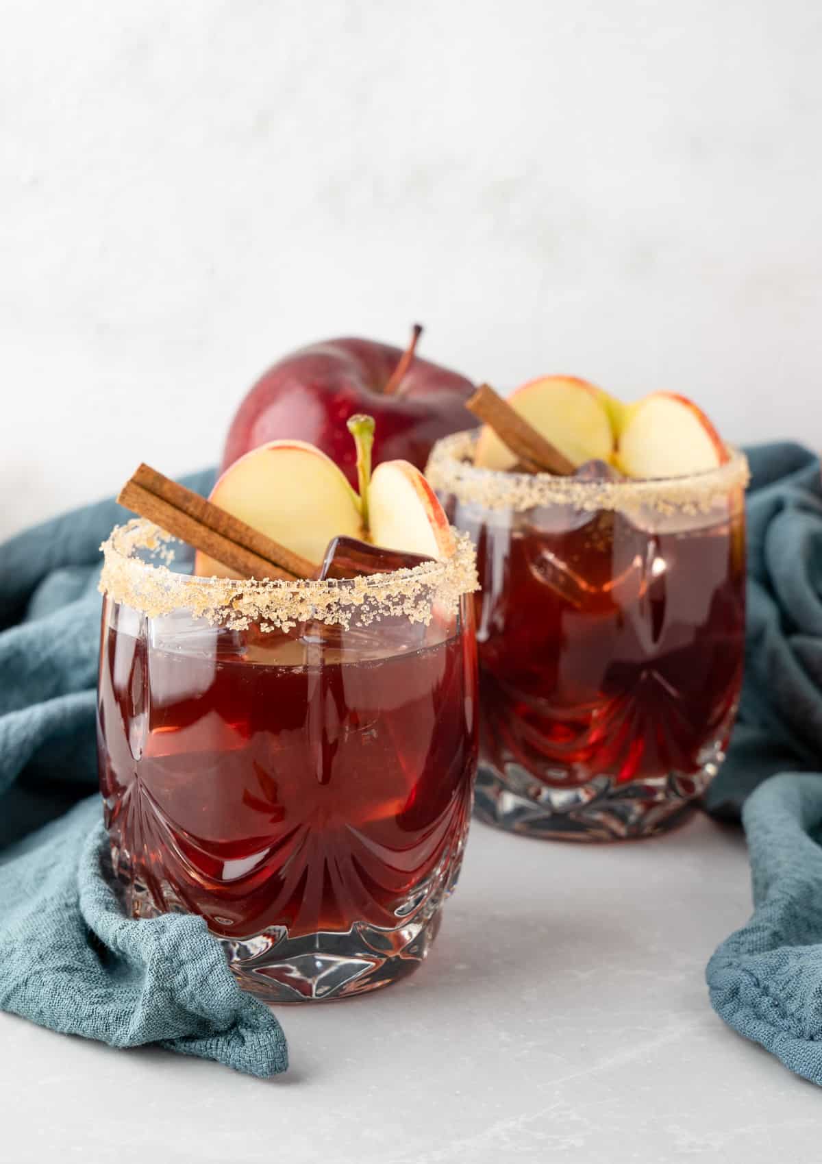 Two glasses of sangria with blue linen garnished with brown sugar rim, sliced apple and cinnamon stick.