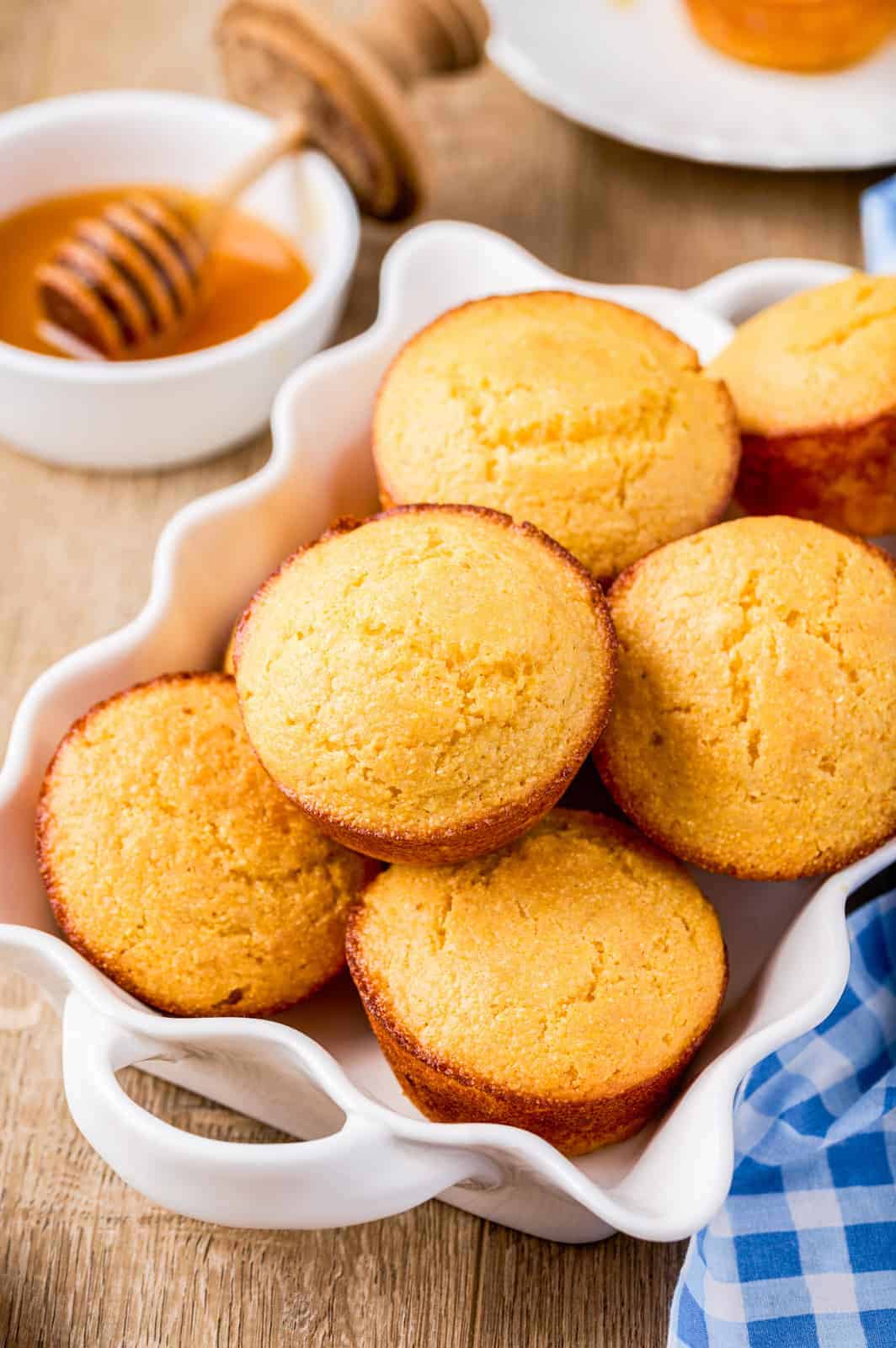 Cornbread Muffins stacked in a scalloped white dish with honey in background.