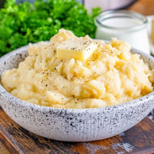 Square image of bowl of mashed potatoes with butter on top and parsley in background.