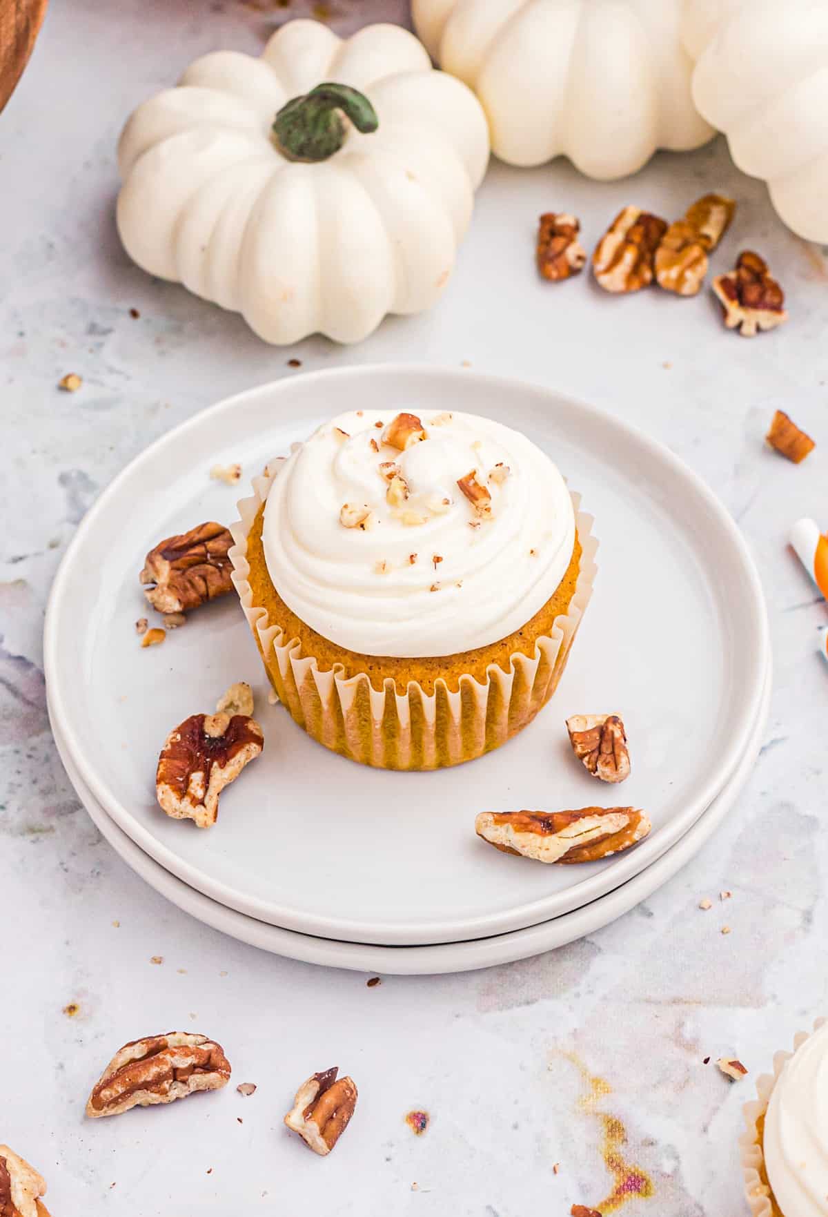 Overhead image of one finished cupcake on white plate with pecans and white pumpkins behind plate.