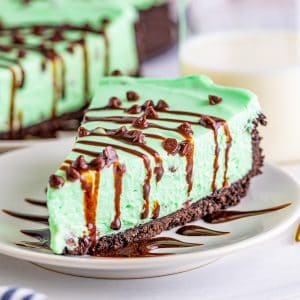 Square image of Mint Cheesecake on white plate with mini chocolate chips drizzled with chocolate syrup.