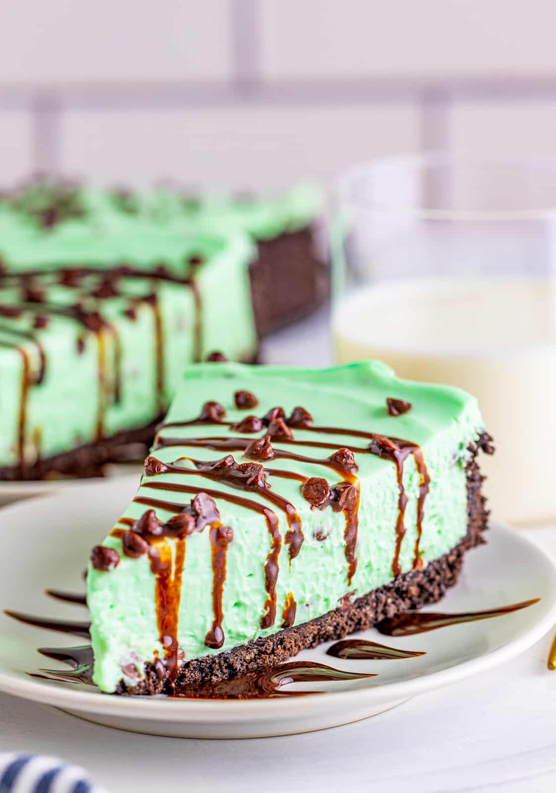 Slice of Mint Chocolate Cheesecake topped with chocolate chips and syrup on white plate.