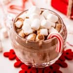 Square image of hot chocolate with marshmallows and candy cane on red snowflake coaster.