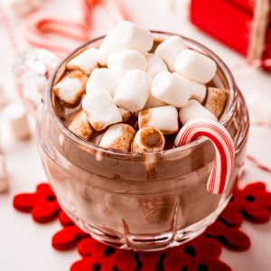 Square image of hot chocolate with marshmallows and candy cane on red snowflake coaster.
