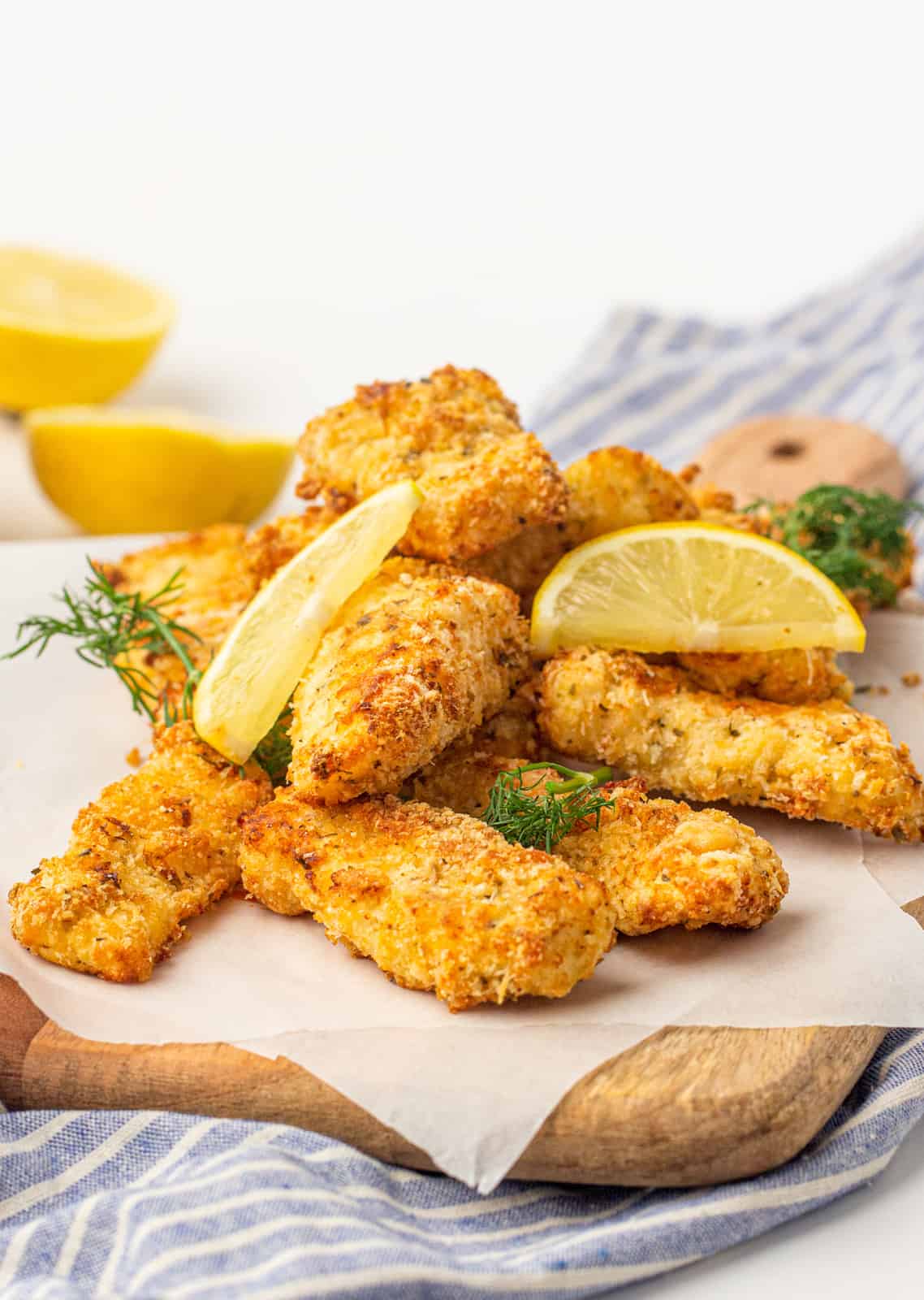 Stacked Air Fryer Fish STicks on cutting board with lemon wedges and dill.