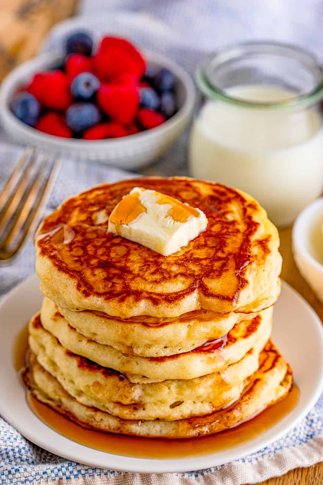 Stacked Buttermilk Pancakes on plate with pat of butter and syrup with fruit in background.