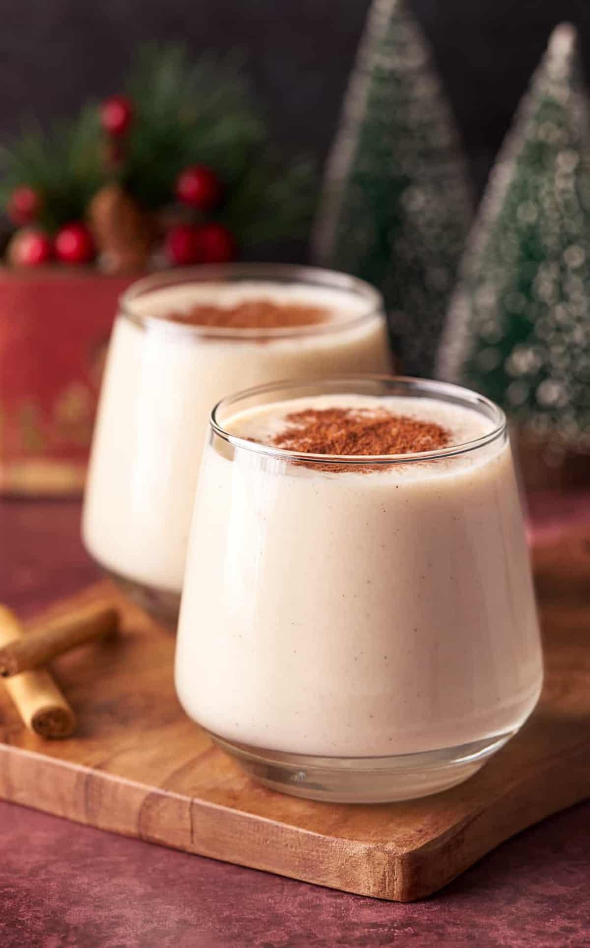 Two glasses of the Coquito on wooden board with cinnamon sticks and topped with ground cinnamon.