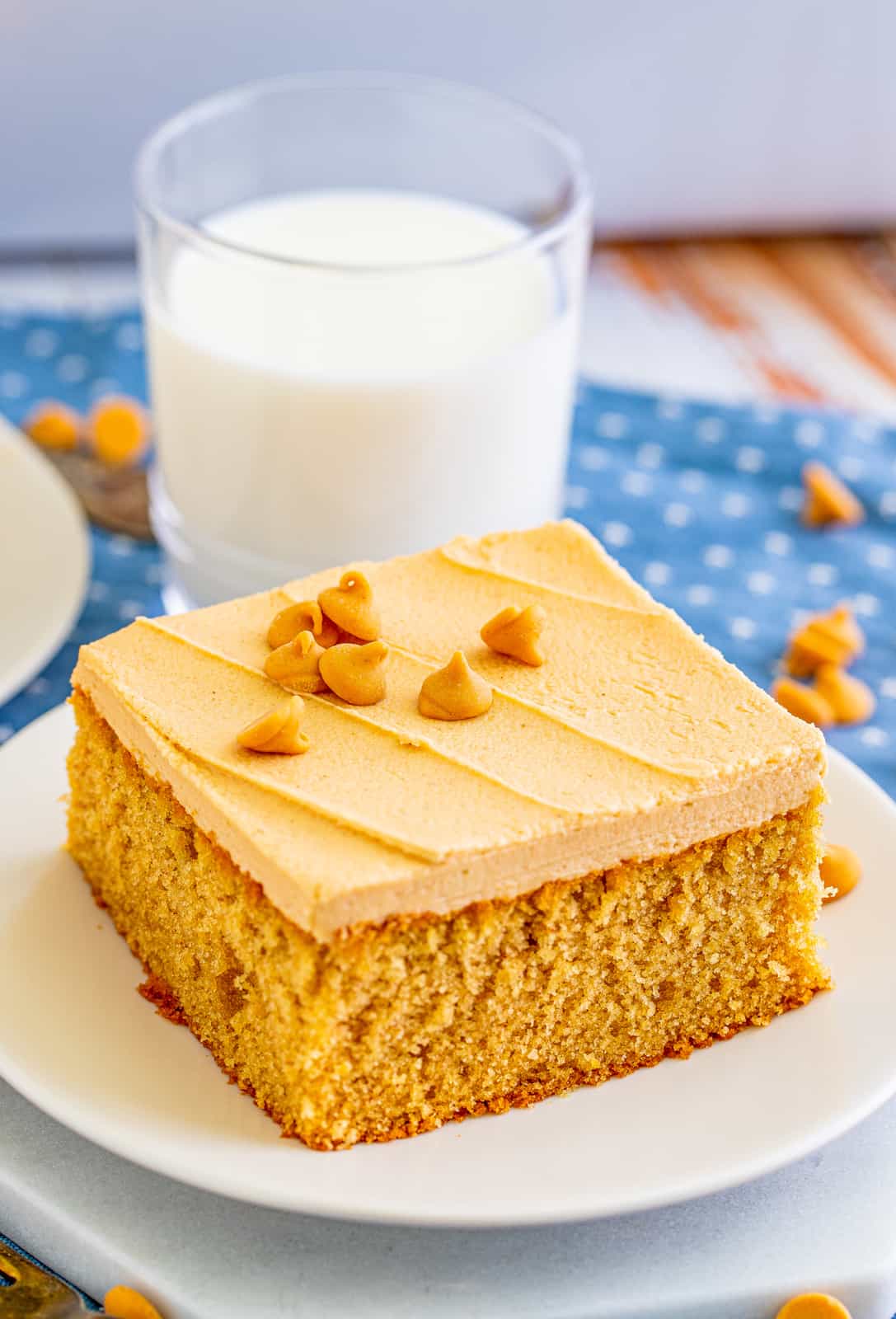 Slice of Peanut Butter Cake on white plate topped with peanut butter chips with milk behind it.