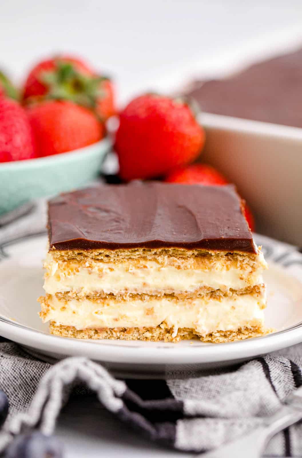 Slice of Eclair Cake on white plate with strawberries behind it.