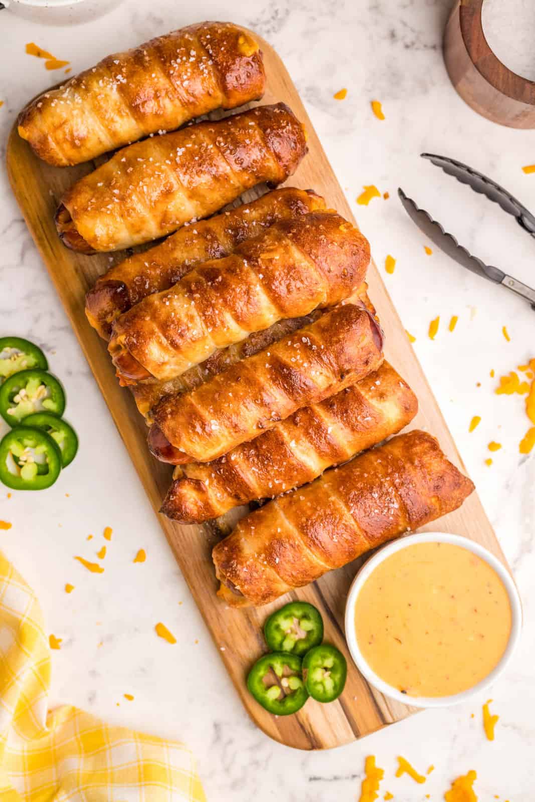 Overhead image of stacked pretzel dogs on wooden board with jalapenos and nacho cheese sauce.