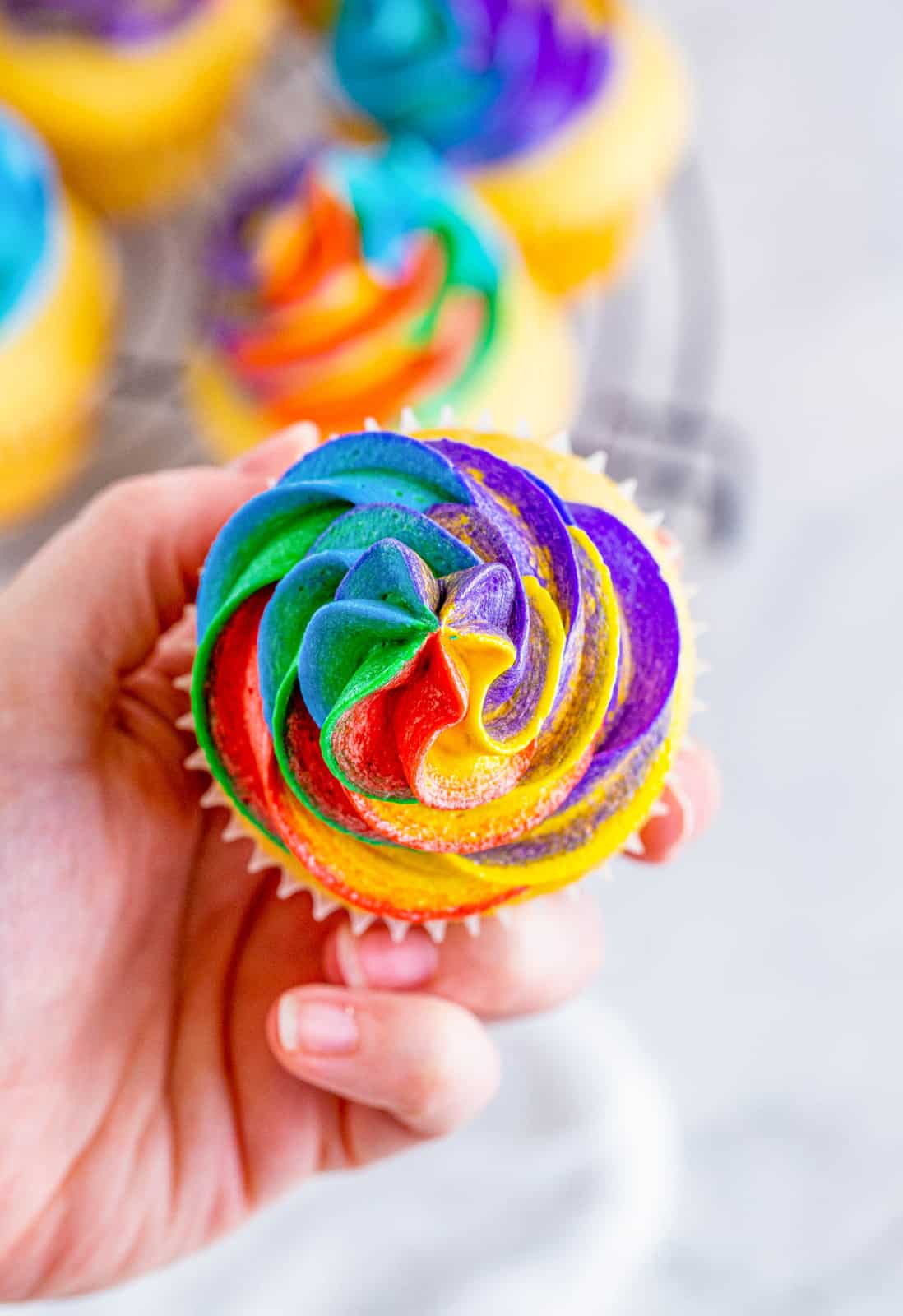 Hand holding up one cupcake showing the top swirls of Rainbow Frosting.