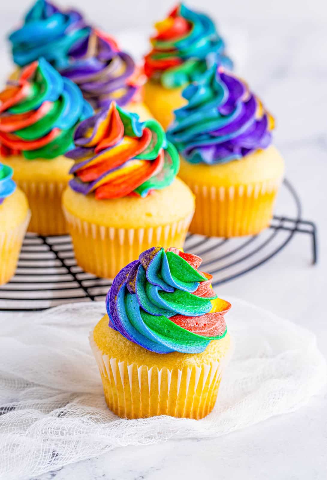 One cupcake frosted with Rainbow Frosting in front of wire rack.
