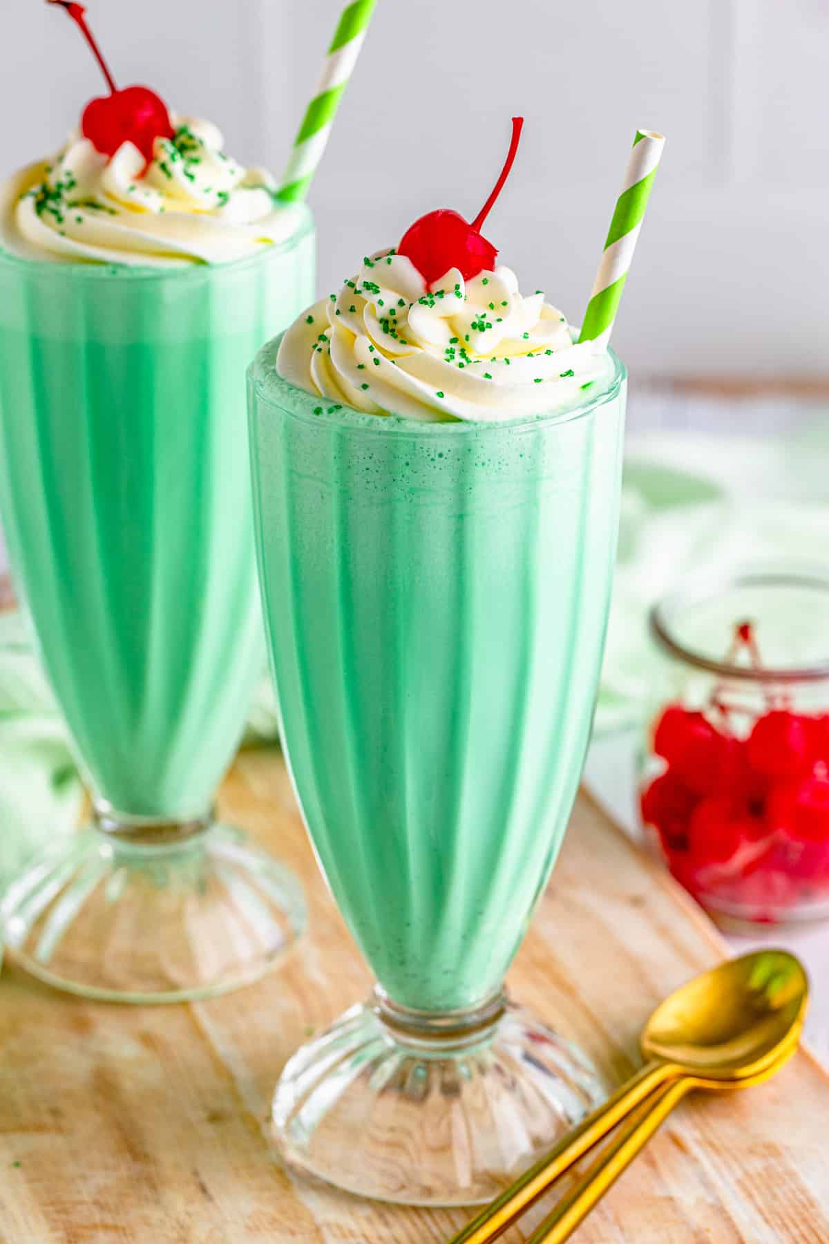 Shamrock Shake Recipe on wooden board with gold spoon. Topped with whipped cream, cheery and green sprinkles.