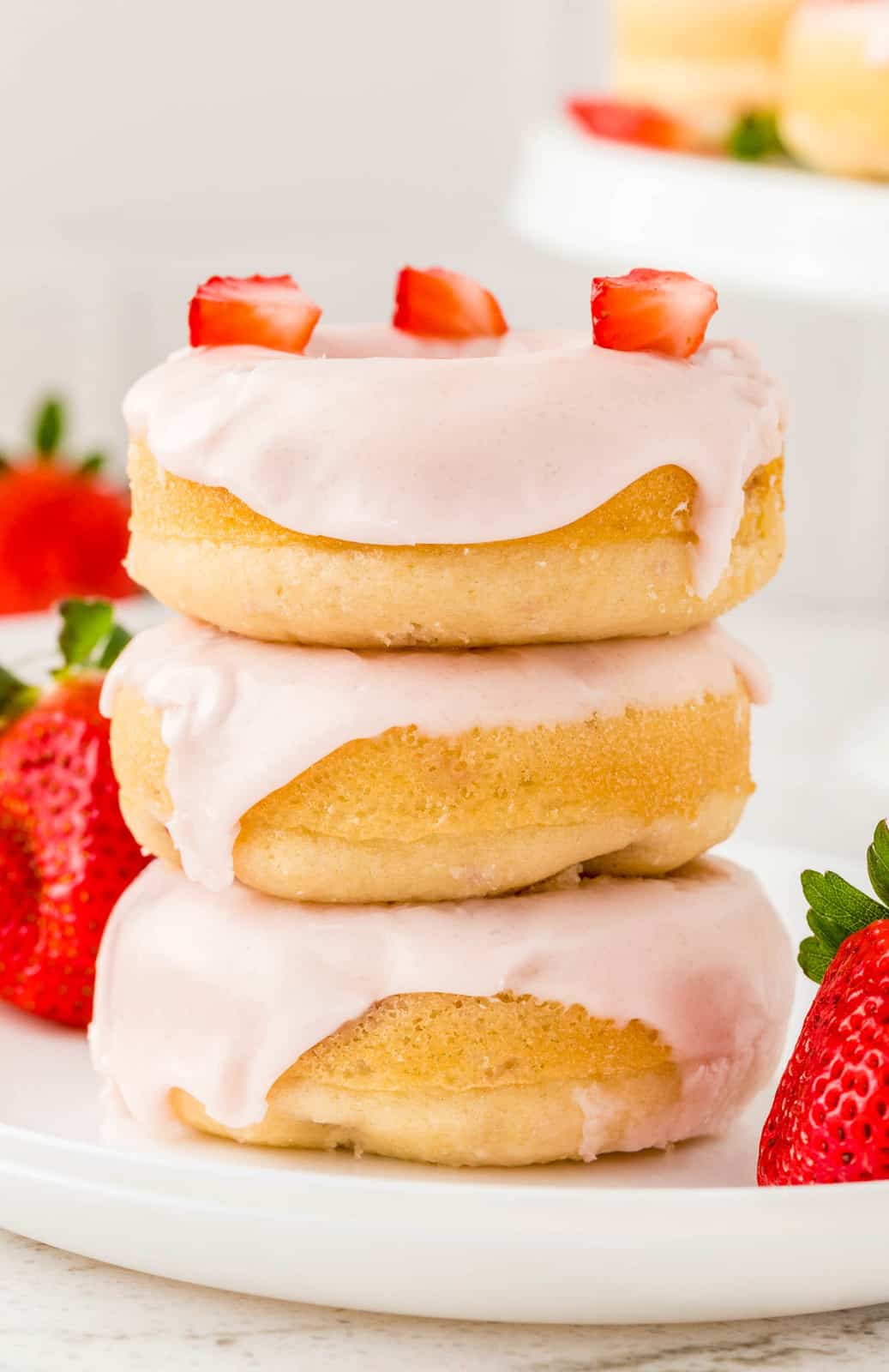 Three stacked Strawberry Donuts on white plate with top donut with strawberries on it.